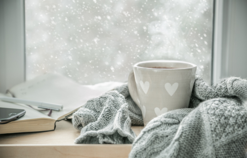 Image is of a cup of coffee, blanket, and notebook in front of a window that shows a snowy landscape, concept of guide to winterize your windows and doors