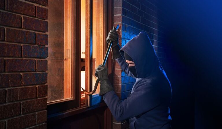 Image is of a burglar using a crow bar to try to break into a house through the window concept of windows and doors impact on home security