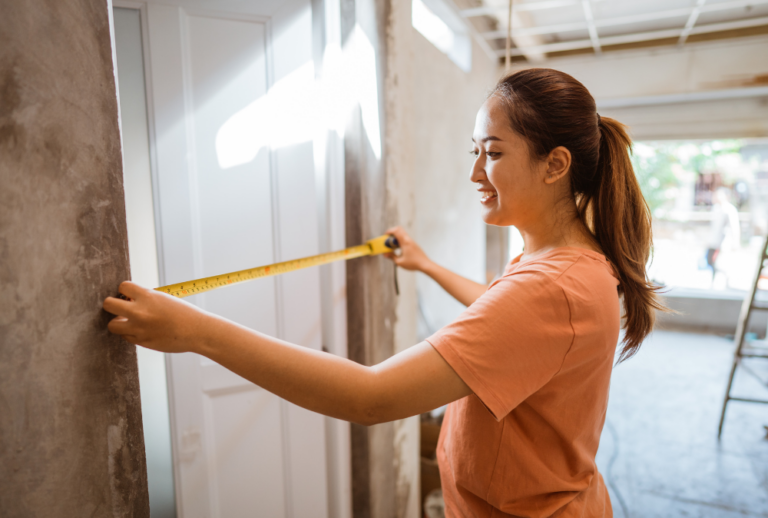 Image is of a woman using a measuring tape to measure for a new door.
