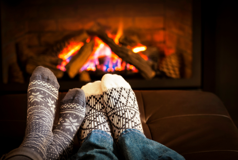 Image is of a couple's feet snuggled up together on a couch in front of a fire, concept of winter-proofing a home, windows, and doors.