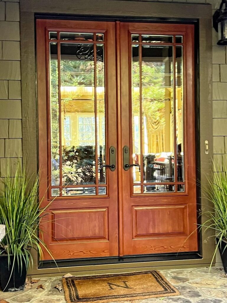 Image is of a set of warm toned wood look 8 foot doors at the entry of a home. The doors have potted grasses on each side and a welcome mat.